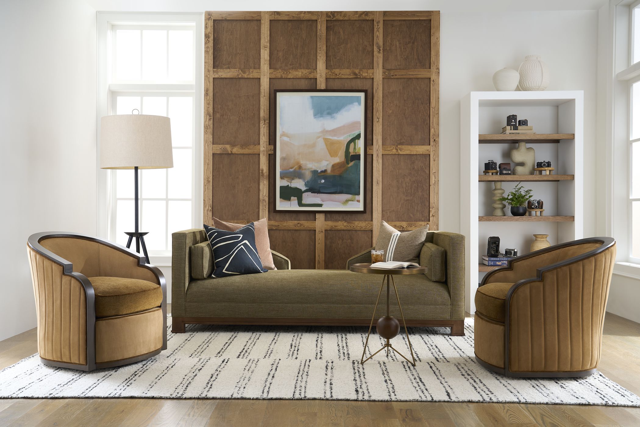 Warm living room with an olive daybed, tan wood-framed swivel chairs, and layered art against a paneled wall