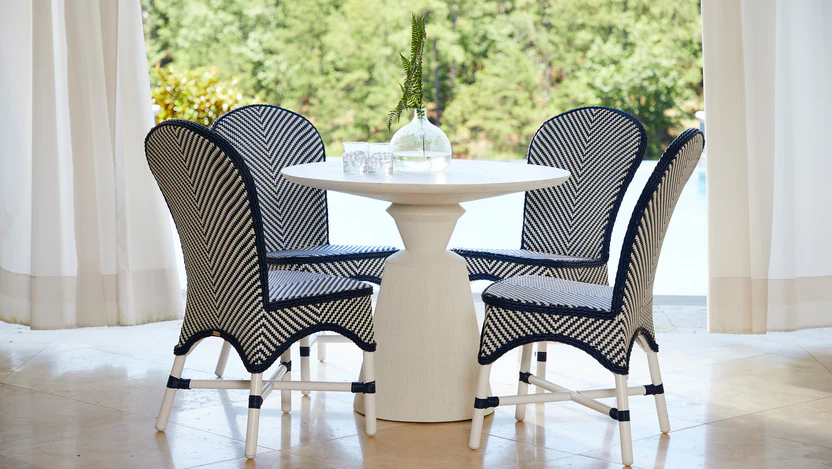 Sunroom dining set with four navy-and-white woven chairs around a round white pedestal table framed by sheer curtains and a garden view