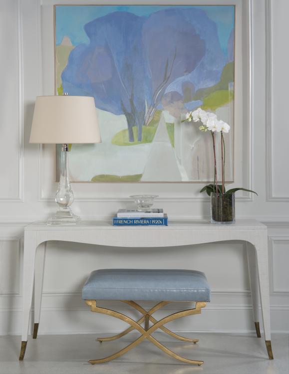 Styled console vignette with a glass table lamp, blue abstract art, orchids, stacked books, and a leather bench